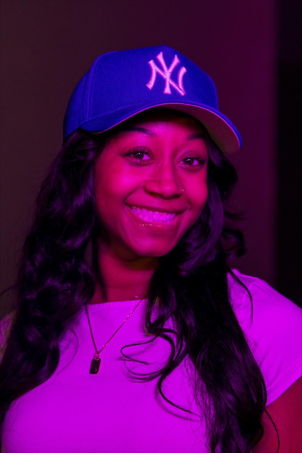 Chrissyyy smiling in a baseball cap under purple lighting