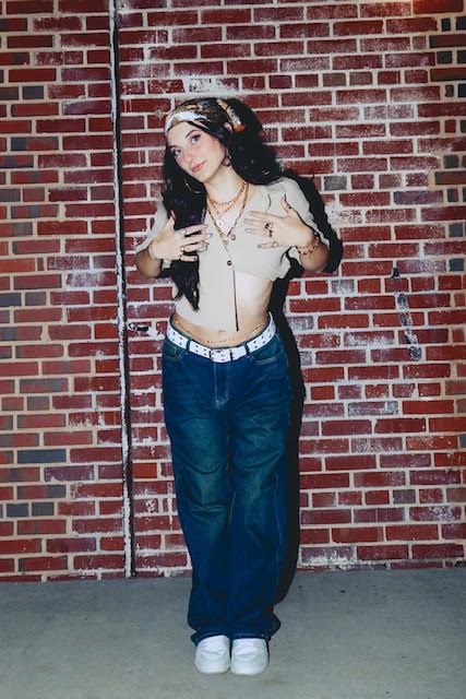 Cosmos posing in a light top and jeans in front of a brick wall